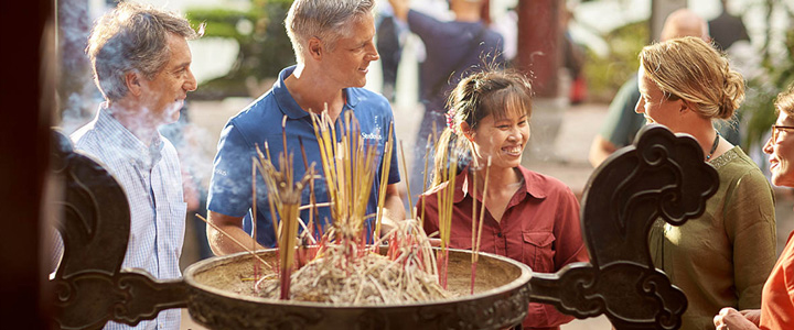 Ein Studiosus-Reiseleiter erläutert zusammen mit einer Vietnamesin seinen Gästen vor einem Behälter mit rauchenden Räucherstäbchen deren Bedeutung.